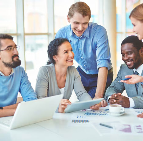 Group of five professionals smiling and discussing work around a table with laptops, charts, and coffee in a bright office setting